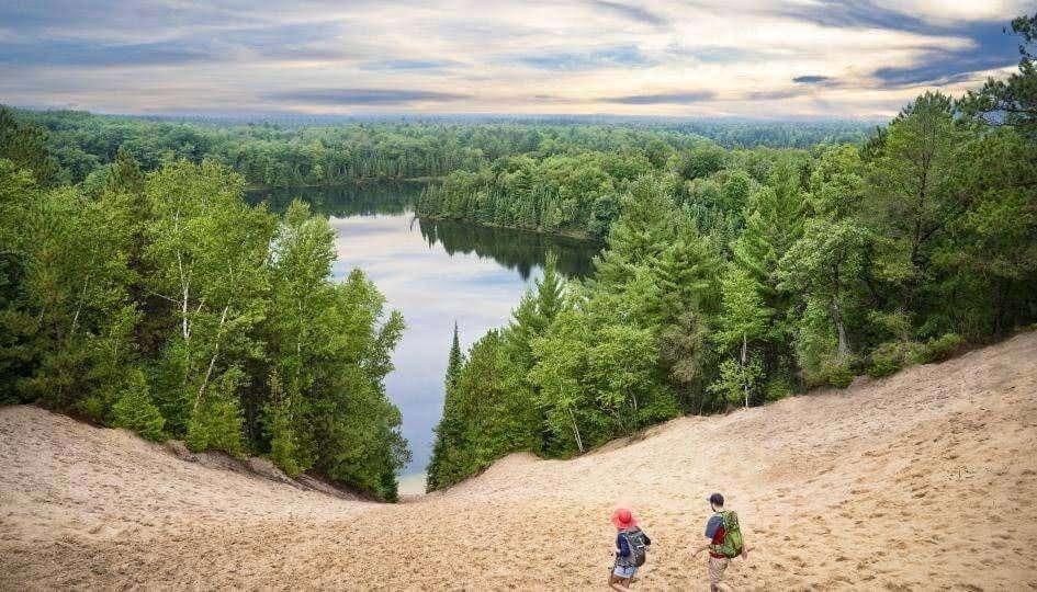 Two hikers walking down a sandy dune surrounded by dense green forest, overlooking a calm lake under a cloudy sky.